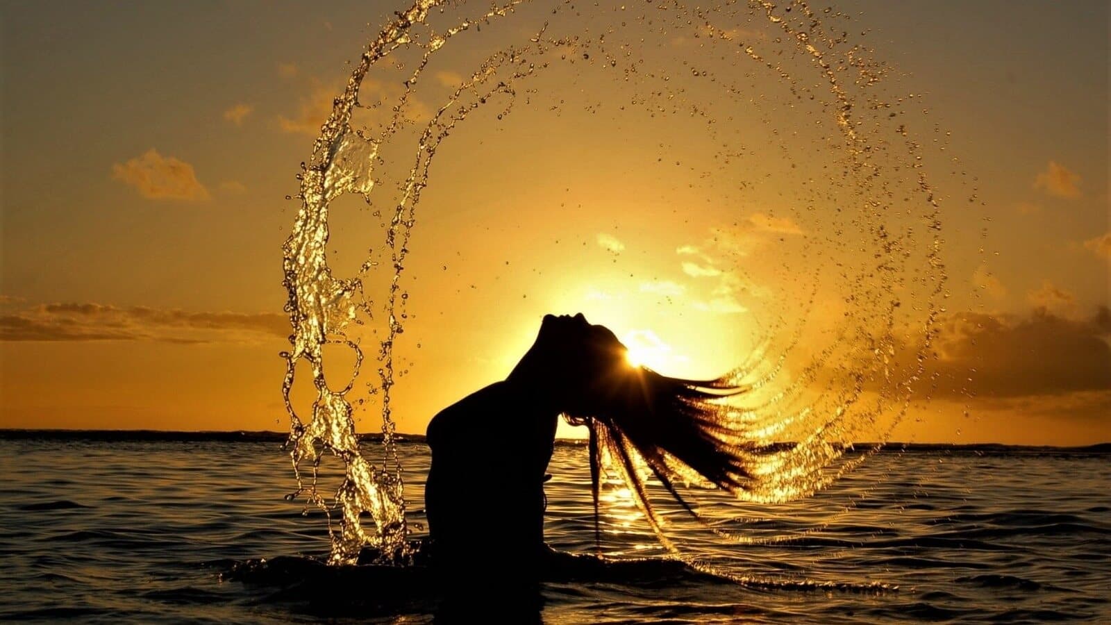 Eine Frau im Meer bei goldenem Sonnenuntergang, das Wasser sprüht im Licht