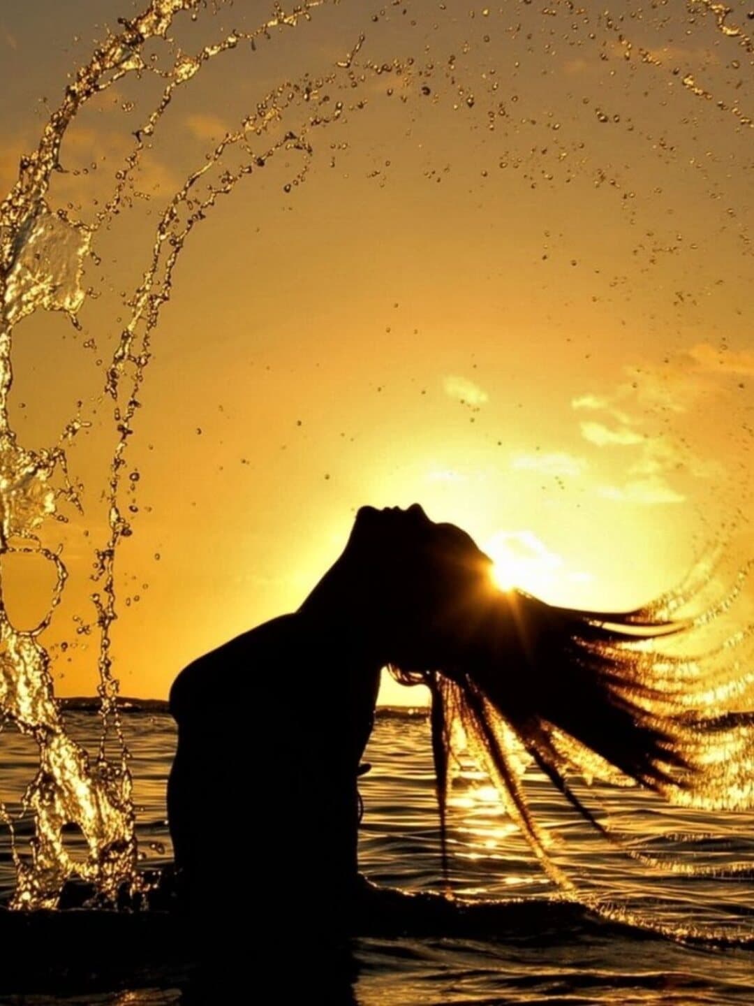Eine Frau im Meer bei goldenem Sonnenuntergang, das Wasser sprüht im Licht
